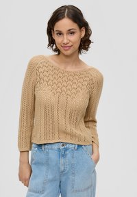 Beige gestrickter Pullover mit offenen und gerippten Mustern, weitem Ausschnitt und dreiviertel Ärmeln, kombiniert mit hellblauen hoch geschnittenen Jeans.