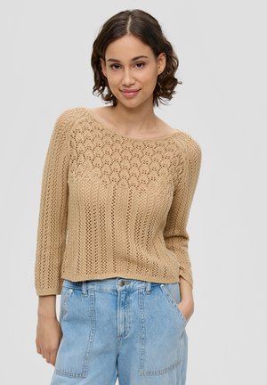 Beige gebreide trui met open en geribbelde patronen, wijde halslijn en driekwartmouwen, gecombineerd met lichtblauwe high-waisted jeans.