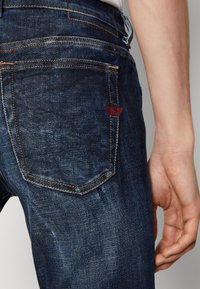 Jeans de mezclilla azul oscuro con costuras naranjas y un logo bordado rojo en el bolsillo trasero. La tela tiene un acabado texturizado.