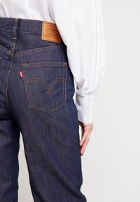 Tmavě modré džíny z denimu s kontrastní koženou nášivkou v barvě tan na pase a dvěma zadními kapsami s charakteristickým vzorem stehů.