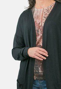 Cardigan negro de material suave con abertura frontal, puños acanalados y dos bolsillos. Lleva encima de una blusa con estampado de flores.