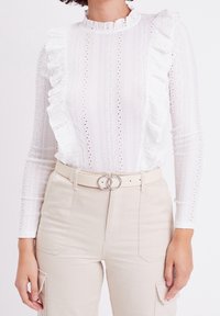 Hvid langærmet bluse med flæse-detaljer og øjenlidsstruktur, parret med beige high-waisted bukser og et rundt bælte.