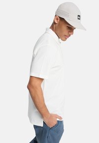 Νεαρός άνδρας που φοράει λευκό πόλο πουκάμισο, ανοιχτό γκρι καπέλο με λογότυπο και μπλε παντελόνι, με τα χέρια στις τσέπες, στραμμένος στο πλάι σε απλό φόντο.