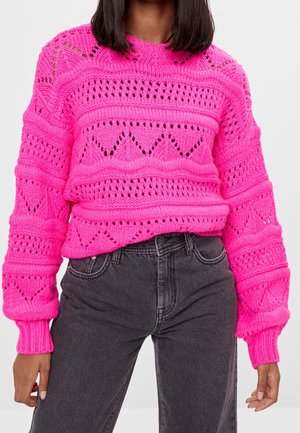 Femme portant un pull en tricot rose vif avec des motifs géométriques associé à un jean taille haute gris foncé, debout devant un fond uni.