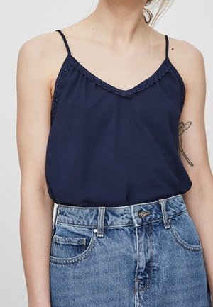 Vrouw draagt een marineblauw topje met kanten rand, gestopt in een high-waist lichtblauwe spijkerbroek, met een zichtbare tatoeage op haar linkerarm.