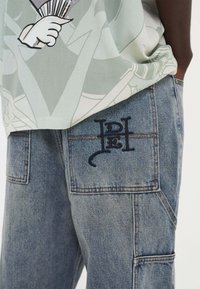 Green graphic T-shirt with abstract design, paired with blue denim cargo pants featuring embroidered black logo on the back pocket.