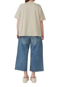 Luźna beżowa koszula z krótkim rękawem, zestawiona z szerokimi niebieskimi dżinsami. Spodnie mają postrzępiony brzeg i tylne kieszenie. Masywne beżowe sandały.