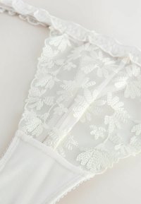 White lace thong featuring floral embroidery, soft mesh, a ruffled waistband, and smooth fabric on the front with scalloped edges.
