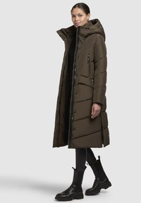 Lungo cappotto imbottito di colore verde oliva con cappuccio, dotato di chiusura a zip, tasche laterali e motivo trapuntato. Abbinato a stivaletti neri alla caviglia.