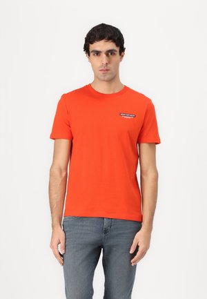 Orange bomull T-skjorte med rund hals, korte ermer, slim fit, med et lite svart merke på brystet. Kombinert med blå jeans.