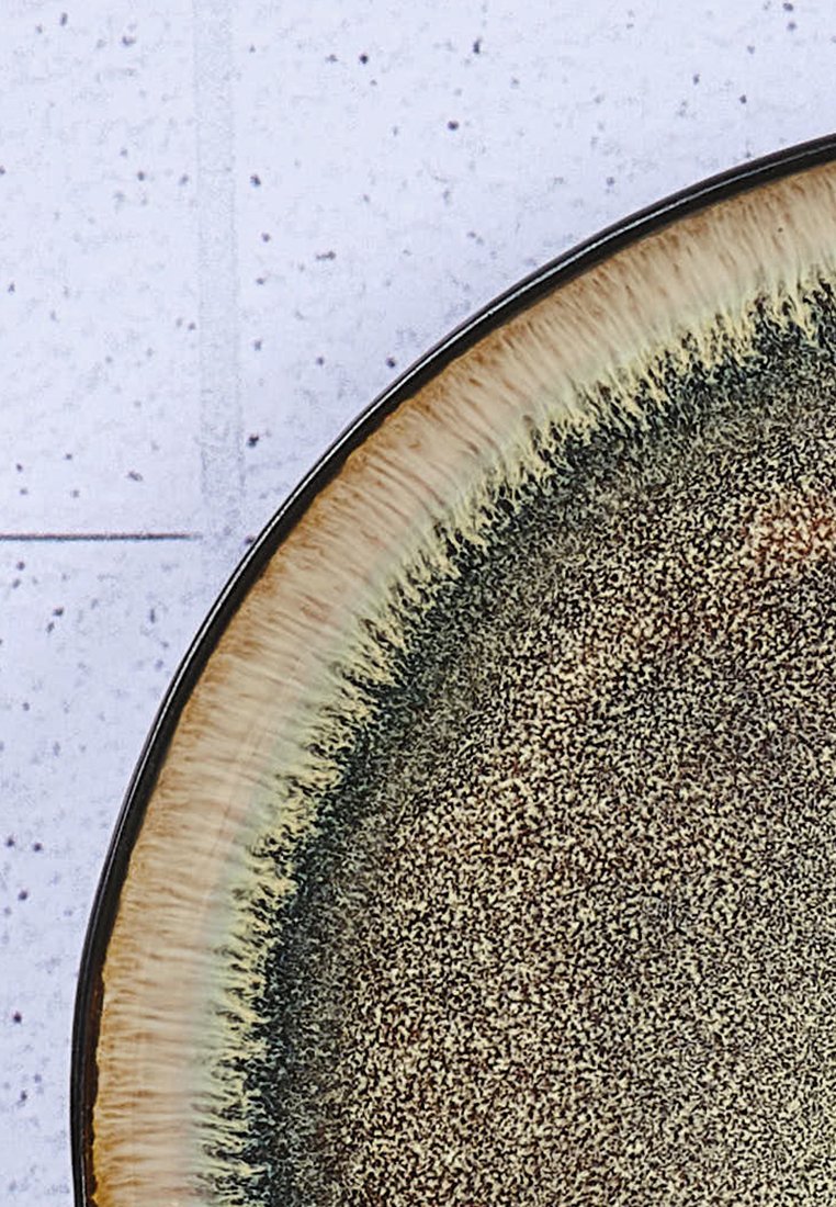 Okrągły ceramiczny talerz o teksturowanej, nakrapianej powierzchni w ziemistych odcieniach. Krawędzie są ciemnobrązowe z gradientem przechodzącym w jaśniejszy beż.