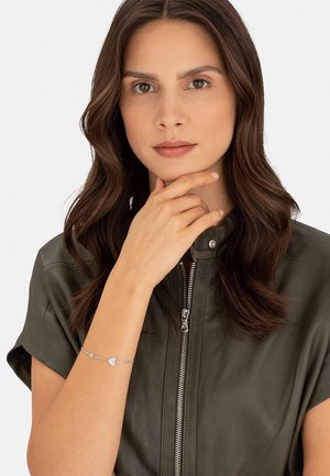 Bracciale d'argento con pendente a forma di cuore e una piccola pietra preziosa, indossato su una mano su uno sfondo di una giacca di pelle verde scuro.
