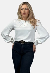 Biała bluzka z balonowymi rękawami, z dziurką przy dekolcie. W zestawieniu z ciemnoniebieskimi dżinsami i czarnym paskiem z klamrą z logo.