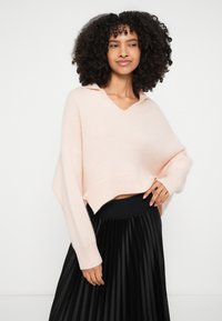 Ανοιχτό ροζ κοντό πλεκτό πουλόβερ με V-λαιμόκοψη και φαρδιά μανίκια, συνδυασμένο με μαύρη πλισέ φούστα. Απαλό ύφασμα, χαλαρή γραμμή.