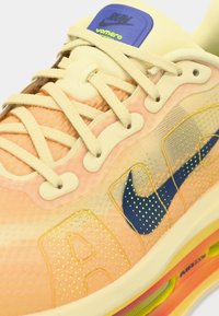 Αθλητικά παπούτσια Nike με διαφανές ροδακινί επάνω μέρος, κίτρινες λεπτομέρειες, μπλε λογότυπο και υφασμένη διακόσμηση. Διαθέτουν μπεζ κορδόνια και μαξιλαρωμένη σόλα.