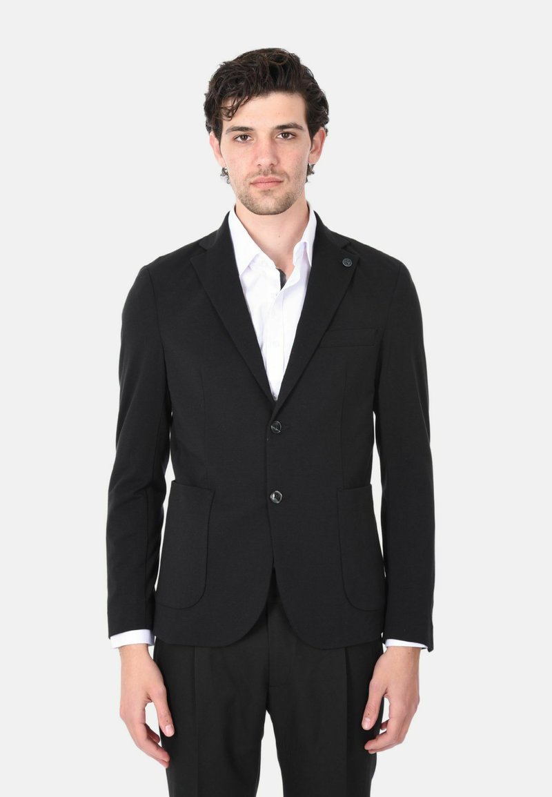 Blazer nero realizzato in tessuto elasticizzato con design aderente, due tasche frontali, chiusura con un bottone e una camicia bianca sotto.