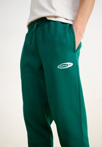 Zelené bavlnené tepláky s elastickým pásom, bočnými vreckami a bielym logom Reebok na ľavom stehne. Mäkkučká textúra a voľný strih.