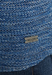 Blaues gestricktes Material mit einem strukturierten Muster, das am unteren Rand ein kleines Metallschild mit der Aufschrift "ONLY & SONS" trägt.