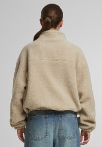 Pullover beige in pile con colletto alto, superficie testurizzata e vestibilità morbida. Abbinato a jeans a vita alta di colore blu chiaro con tasche posteriori.