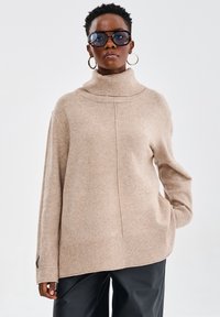 Beżowy sweter z golfem, z ribbowanym dołem i mankietami, o luźnym kroju i teksturowanej tkaninie. Stylizowany z czarnymi spodniami o szerokich nogawkach.