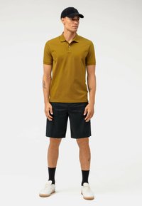 Polo verde oliva con colletto classico, abbinato a pantaloni corti neri. Il modello indossa sneakers bianche e un cappellino nero. Design minimale.