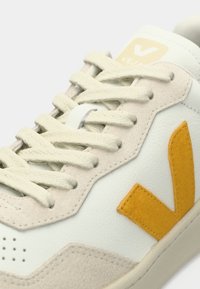 Baskets blanches et beiges avec des lacets crème, un logo "V" jaune sur le côté, et la marque "Veja" sur la languette.