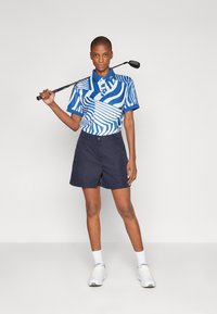 Polo shirt da golf blu e bianco con motivi a righe orizzontali e verticali, abbinato a pantaloni corti blu scuro e scarpe da ginnastica bianche.