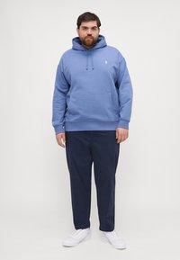 Sudadera azul con bolsillo frontal, capucha con cordones y pequeño logo. Combinada con pantalones marino y zapatillas blancas. Sin patrones visibles.