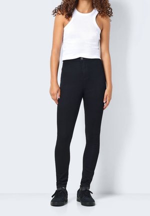 Femme aux cheveux bouclés portant un haut à rayures sans manches blanc, un pantalon skinny noir taille haute et des baskets noires, se tenant devant un fond uni.