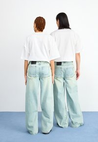Two people wearing oversized white t-shirts and light blue wide-leg jeans with frayed hems, standing back-to-back on a blue surface.