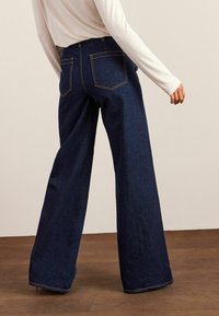 Personne portant un jean en denim large bleu foncé et un haut à manches longues beige clair, se tenant sur un sol en bois contre un mur neutre.