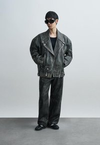 Üle suurte mõõtudega must nahktagi hõbedaste detailidega, kombineeritud mustade pükste ja mustade jalanõudega. Modell kannab julgeid päikeseprille. Tekstuuriga viimistlus.