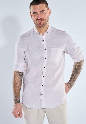 Man with short hair and tattoos wearing a light linen shirt with rolled-up sleeves and light-colored pants, standing against plain background.