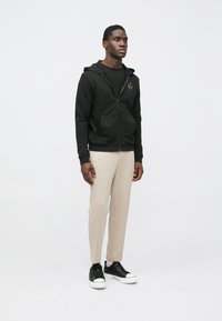 Čierna mikina na zips s prednými vreckami a zlatým logom, doplnená svetlobéžovými zúženými nohavicami a čiernymi teniskami s bielymi podrážkami.