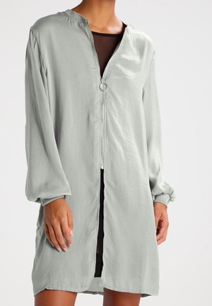 Mujer con vestido de manga larga color gris claro con cremallera frontal y tirador redondo, sobre prenda interior negra.