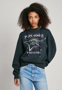 Hoodie noir surdimensionné avec impression graphique présentant un design d'étoile et "Pepe Jeans London". Cordons réglables à la capuche. Porté avec un jean bleu clair.
