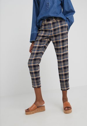 Frau trägt karierte, kurze Hosen und ein blaues Jeanshemd, steht mit Händen in den Taschen und trägt beige Keilsandalen vor weißem Hintergrund.