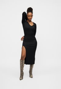 Μαύρο στενό φόρεμα με μακριά μανίκια και πλαϊνή σχισμή, συνδυασμένο με μπότες στιλέτο πάνω από το γόνατο με τύπωμα φιδιού. Λεία υφή υφάσματος και εφαρμοστή γραμμή.