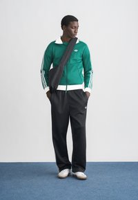 Green athletic jacket with white collar and stripes, black pants, and white shoes. Model poses with a black crossbody bag.