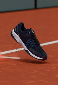 Svart och blå tennissko som sparkar upp lera på en röd lertennisbana nära baslinjen.