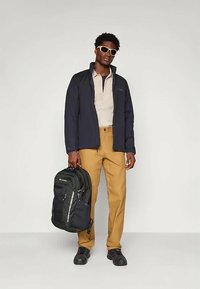 Temno modra jakna, bež srajca, rjave hlače, črna nahrbtnik z zadrgami in temni čevlji; sončna očala dopolnjujejo zunanji videz.