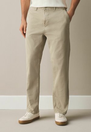 Mann trägt beige legere Hose und weiße Turnschuhe und steht mit den Händen in den Taschen vor einer schlichten Wand.