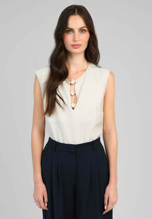 Femme aux longs cheveux foncés portant un haut blanc sans manches avec trois boutons en forme de coquillage et un pantalon noir plissé taille haute.