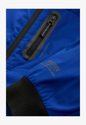 Manica di giacca blu con polsino elastico nero, una tasca con zip verticale con rifiniture nere e un piccolo testo stampato vicino al polsino.