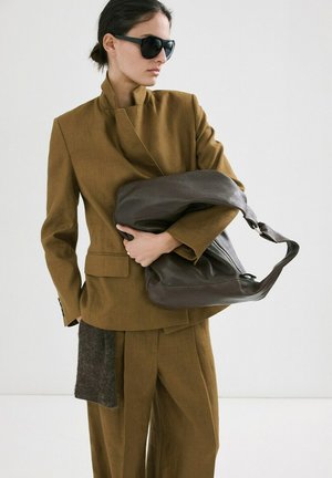 Person in braunem, maßgeschneidertem Anzug und dunkler Sonnenbrille, die eine große, schlaffe dunkle Ledertasche vor einem einfarbigen Hintergrund hält.