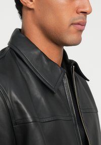 Chaqueta de cuero negra con un colla ancha, que presenta un cierre frontal, textura suave y costuras horizontales en el pecho.