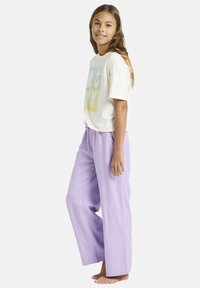 Młoda dziewczyna stoi boso, ubraną w biały t-shirt z grafiką i luźne jasnofioletowe spodnie, uśmiecha się, ma długie brązowe włosy.