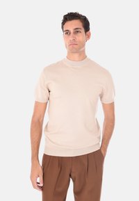 Uomo in piedi con le mani in tasca, che indossa una maglietta a maniche corte in maglia beige e pantaloni marroni a pieghe, su uno sfondo semplice.