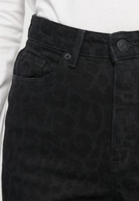 Close-up of black high-waist jeans with subtle leopard print pattern, button closure, and a front pocket worn with a white top.
