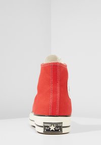 Red canvas high-top sneaker with white stitching and a circular logo. Features a black rubber sole with "Converse All Star" branding.